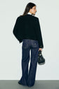 Woman standing facing away, wearing a black jacket, white shirt, and dark blue wide-leg jeans à coupe large, while holding a small black handbag. She has long brown hair and is standing against a plain light background.