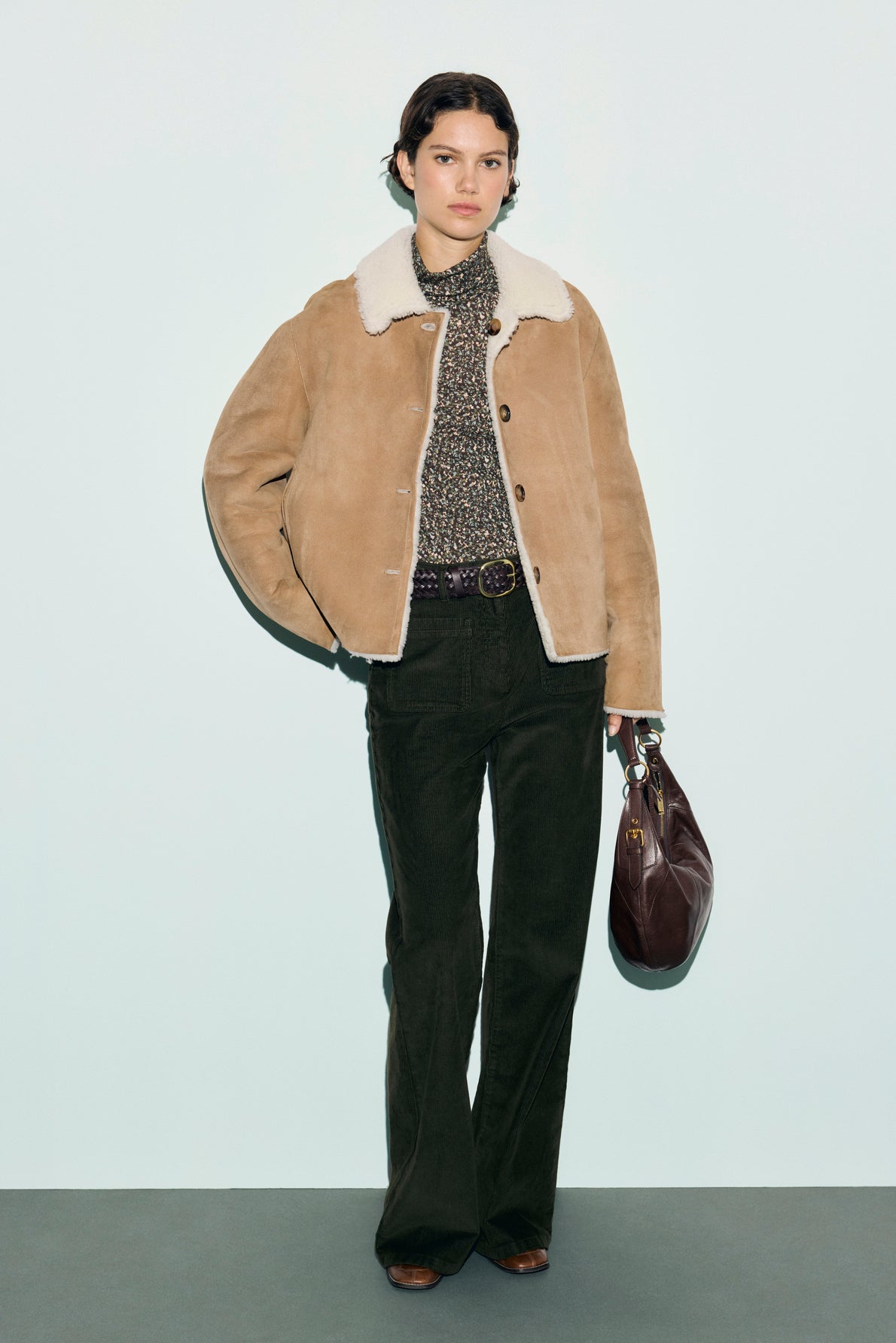 A person wearing a tan shearling jacket, patterned shirt, and pantalon long kaki à coupe évasée stands holding a large brown handbag against a plain light background. Brown shoes complete the look.