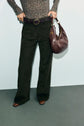 Person wearing a patterned long-sleeve top, dark coupe évasée wide-leg pants, brown belt, and brown shoes, holding a large brown purse against a plain light background. Face is not visible in the photo.