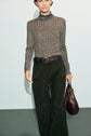 A person wearing a patterned turtleneck top, a pantalon long kaki in dark green corduroy with patch pockets, a braided belt, and holding a brown leather handbag.