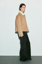A person stands against a plain background wearing a tan shearling jacket with white lining, paired with a pantalon long kaki that adds a touch of sophistication. They complete the look with brown shoes and have short dark hair and a neutral expression, looking slightly to the side.