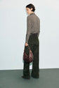 A person with short dark hair is standing sideways, wearing a patterned long-sleeve top and green pantalon long kaki à coupe évasée, holding a brown shoulder bag, against a plain light background.
