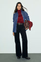 Woman in a red turtleneck, blue denim shirt, black wide-leg pants inspired by the chic look of a pantalon long femme, and black shoes, holding a brown bag and a red cap, stands against a plain background.