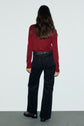 A person with long brown hair stands facing away, wearing a red long-sleeve top, a jean évasé marron, a braided belt, and dark shoes, against a plain light background.