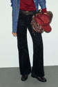Person wearing a red top, blue denim shirt, black wide-leg pants reminiscent of a pantalon long femme, black shoes, and a black belt, holding a brown bag and a red baseball cap. Face is not visible.