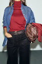 A person wearing a red turtleneck, blue denim shirt, black pants reminiscent of a pantalon long femme style, and a brown belt holds a brown leather bag and a red cap. Their face is not visible.