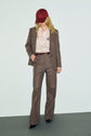 A person wearing a brown suit, striped shirt, dark belt, and a red cap stands against a plain background, looking down with hands in pockets—an effortlessly cool look bureau chic that pairs perfectly with their tailored pantalon de tailleur.