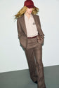 A person wearing a brown suit and pantalon de tailleur, paired with a striped shirt, maroon belt, and maroon cap, is walking with hands in pockets against a plain background, embodying a look bureau chic.