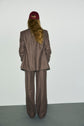 A person with long hair wearing a burgundy cap and an oversized brown pantalon de tailleur stands facing a white wall, with their back to the camera, embodying a look bureau chic.