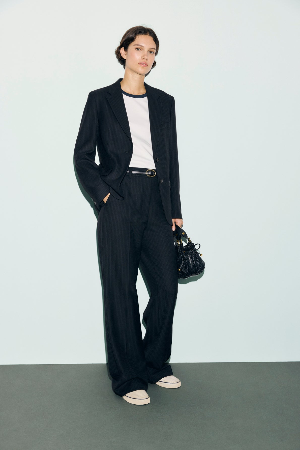 A person wearing a black pinstripe suit with a pantalon de tailleur, white t-shirt, black belt, and white sneakers stands against a light background, holding a small black handbag.