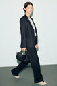 A person with short dark hair wears a black pantalon de tailleur suit, a white T-shirt, and sneakers while walking and holding a black handbag against a plain light background.