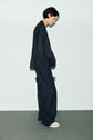 Person with short dark hair stands sideways wearing an oversized black suit, featuring a pantalon de tailleur, and white shoes against a pale background. Their hands are in their pockets, and they have a calm, relaxed posture.