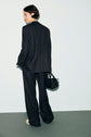 A person with short dark hair, dressed in a black suit and pantalon de tailleur, stands facing away. They hold a black woven handbag and wear light-colored shoes, with a plain background behind them.