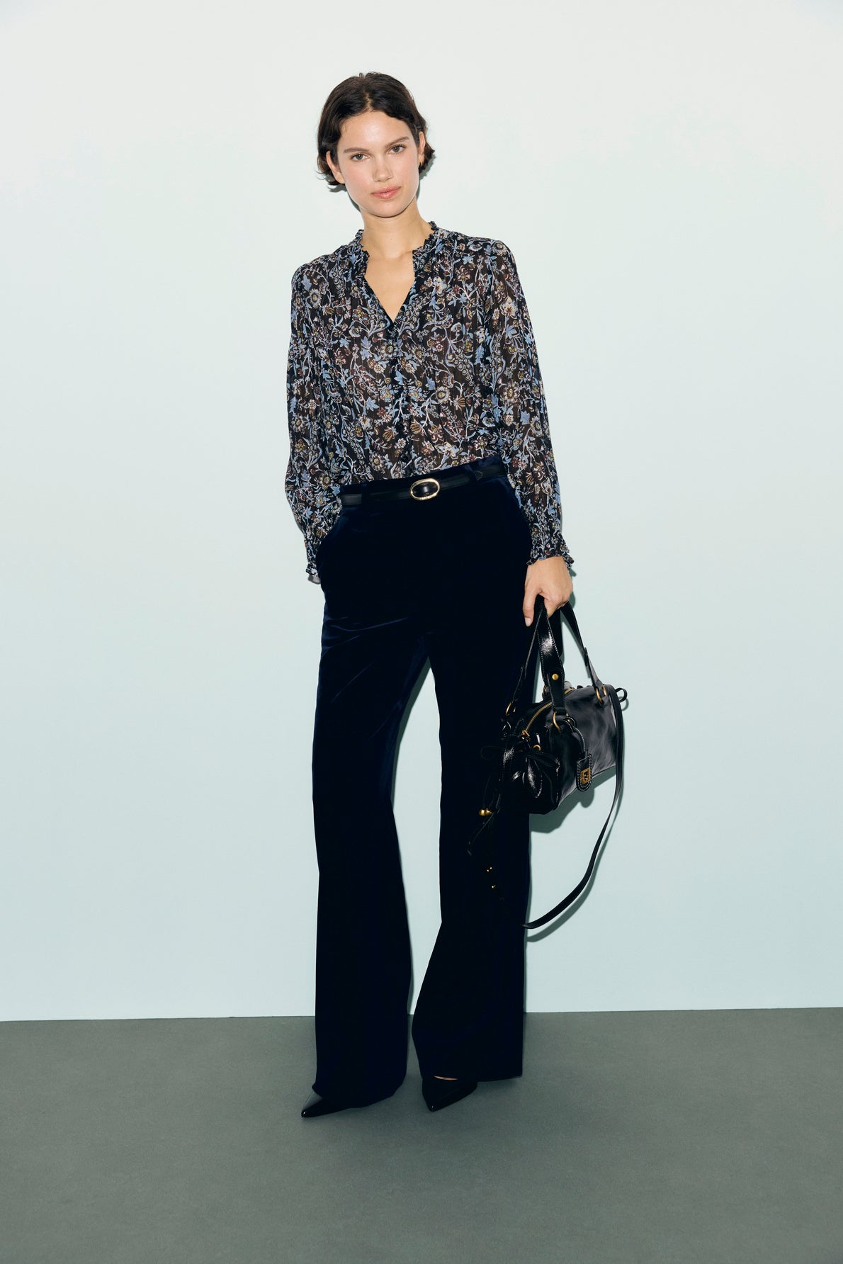 Woman standing against a light background, wearing a patterned blouse, a chic pantalon tailleur femme in dark flared style, and holding a black handbag.