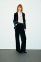 A person with wavy, reddish hair wears a dark velvet suit, featuring a pantalon tailleur velours paired with a light button-up shirt and black shoes, standing against a plain light background.