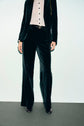 A person wearing a black pantalon tailleur velours suit with flared pants, a white button-up shirt, and black shoes stands against a plain light background. Their head is not visible in the image.