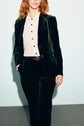A person wearing a dark velvet suit with a white button-up shirt and a black belt stands against a plain background, one hand in their pocket and a small purse on their shoulder, showcasing an elegant pantalon tailleur velours.