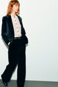 A person with shoulder-length red hair wearing glasses, a dark velvet suit and matching pantalon tailleur velours, a light button-up shirt, and black shoes walks with hands in pockets against a plain white wall.