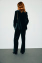 Person with wavy, shoulder-length hair wearing a dark pantalon tailleur velours and holding a bag with a chain strap, standing with their back to the camera against a plain background.