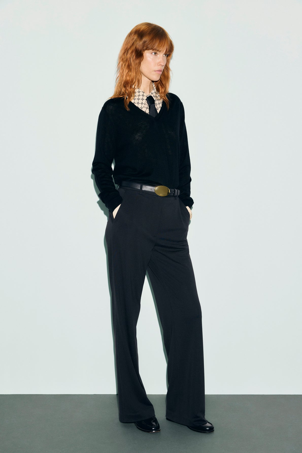 A person with wavy red hair stands against a plain background, wearing a black jersey noir sweater over a patterned shirt, black pantalon tailleur wide-leg trousers, a belt, and black shoes, hands in pockets, looking to the side.