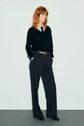 A person with wavy red hair stands against a plain background, wearing a black jersey noir sweater over a patterned shirt, black pantalon tailleur wide-leg trousers, a belt, and black shoes, hands in pockets, looking to the side.