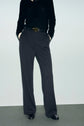 Person wearing a black long-sleeve jersey noir top, black belted pantalon fluide wide-leg trousers, and black shoes stands with hands in pockets against a plain background.