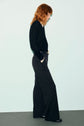 A person with red hair, wearing a black sweater in jersey noir and wide-leg pantalon fluide, stands in profile with hands in pockets against a plain, light background.