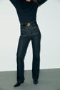A person wearing a long-sleeved black top, a jean droit in denim noir chiné with grandes poches à boutons dorés on the front, and black pointed shoes stands against a plain light-colored wall. Their face and arms are not visible.