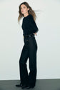 A person with long brown hair stands sideways against a plain background, wearing a black long-sleeve top, a jean droit in denim noir chiné featuring grandes poches à boutons dorés on the front, and black shoes.
