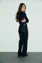 A person with long hair, wearing a black long-sleeve top and dark jeans droit with grandes poches à boutons dorés, stands on a gray floor against a plain white wall, smiling and looking over their shoulder with arms crossed.