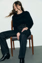 A person in a black long-sleeve shirt, denim noir chiné jean droit with grandes poches à boutons dorés, and black boots sits casually on a wooden chair, hair flowing sideways, against a plain white wall and gray floor.