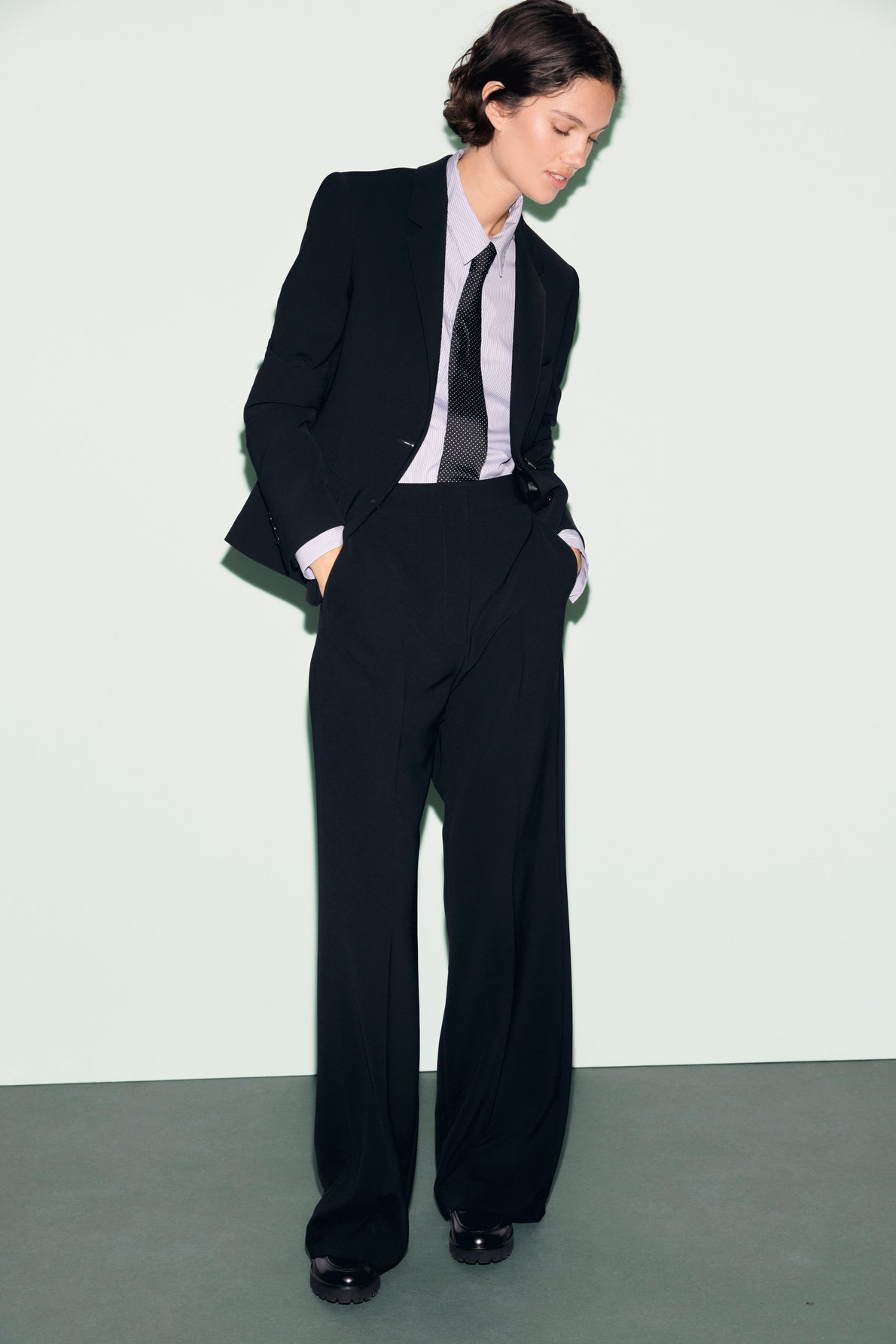 Person wearing a black suit with wide-leg trousers, specifically a pantalon tailleur noir, a light gray shirt, and a loose black tie stands against a pale background with hands in pockets, looking down.