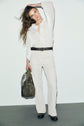 A woman in a white blouse and white pants stands against a plain wall, holding a green handbag. Showcasing a casual chic look, she wears a dark belt and black shoes, with her long hair draped over one shoulder. Her pants feature une coupe droite for an effortlessly stylish touch.