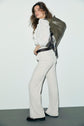 A woman in a white blouse and light pants stands sideways, looking at the camera. She carries an olive-green bag and wears black shoes, embodying a casual chic style. The coupe droite of her pantalon velours côtelé adds a modern touch to the overall look. The background is plain white.
