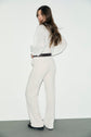 Woman with long brown hair stands sideways, looking over her shoulder. She wears a white blouse, matching coupe droite pantalon velours côtelé, a dark belt, and black shoes against a plain white background for a casual chic look.
