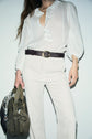 Woman wearing a white blouse and coupe droite white pants in pantalon velours côtelé, styled with a braided dark belt, holding a green handbag with a patterned scarf tied to it. The photo is cropped from the shoulders down for a distinctly casual chic look.