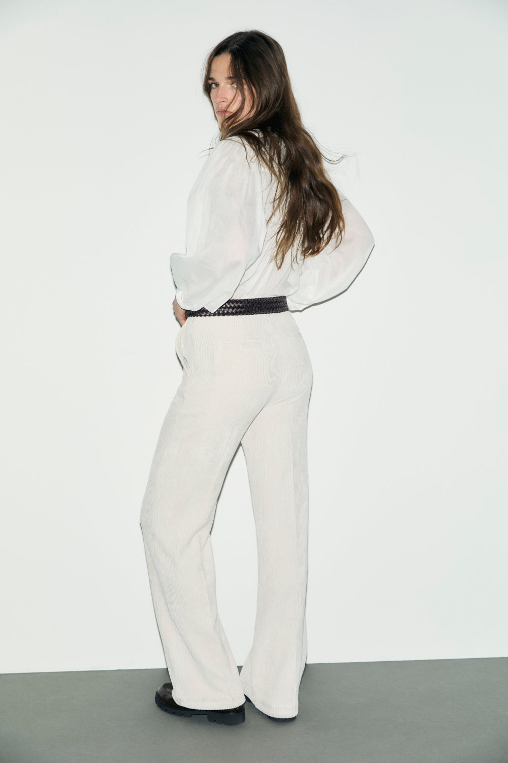 A woman with long brown hair, wearing a white blouse and casual chic pantalon velours côtelé with a dark belt, stands on a gray floor, looking over her shoulder against a white background.