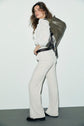A woman in a white blouse and light pants stands sideways, looking at the camera, with a textured green bag over her shoulder. She has loose hair and is wearing black shoes, effortlessly embracing a casual chic style with her coupe droite pantalon velours côtelé.