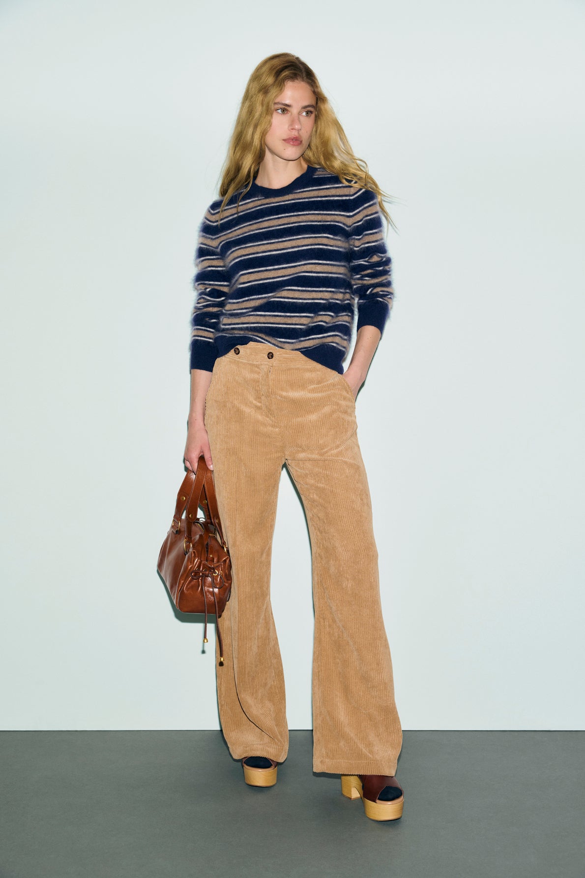 Woman with long blonde hair wearing a navy striped sweater and a beige pantalon velours côtelé, holding a brown handbag and wearing platform shoes, stands against a light background.