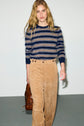A person with long blonde hair wears a navy and beige striped sweater and a pantalon velours côtelé in tan, holding a brown bag, standing against a plain light background.