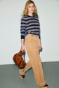 A person wearing a navy and beige striped sweater, a pantalon velours côtelé in a tan shade, and clogs carries a brown leather bag while walking against a plain light background.