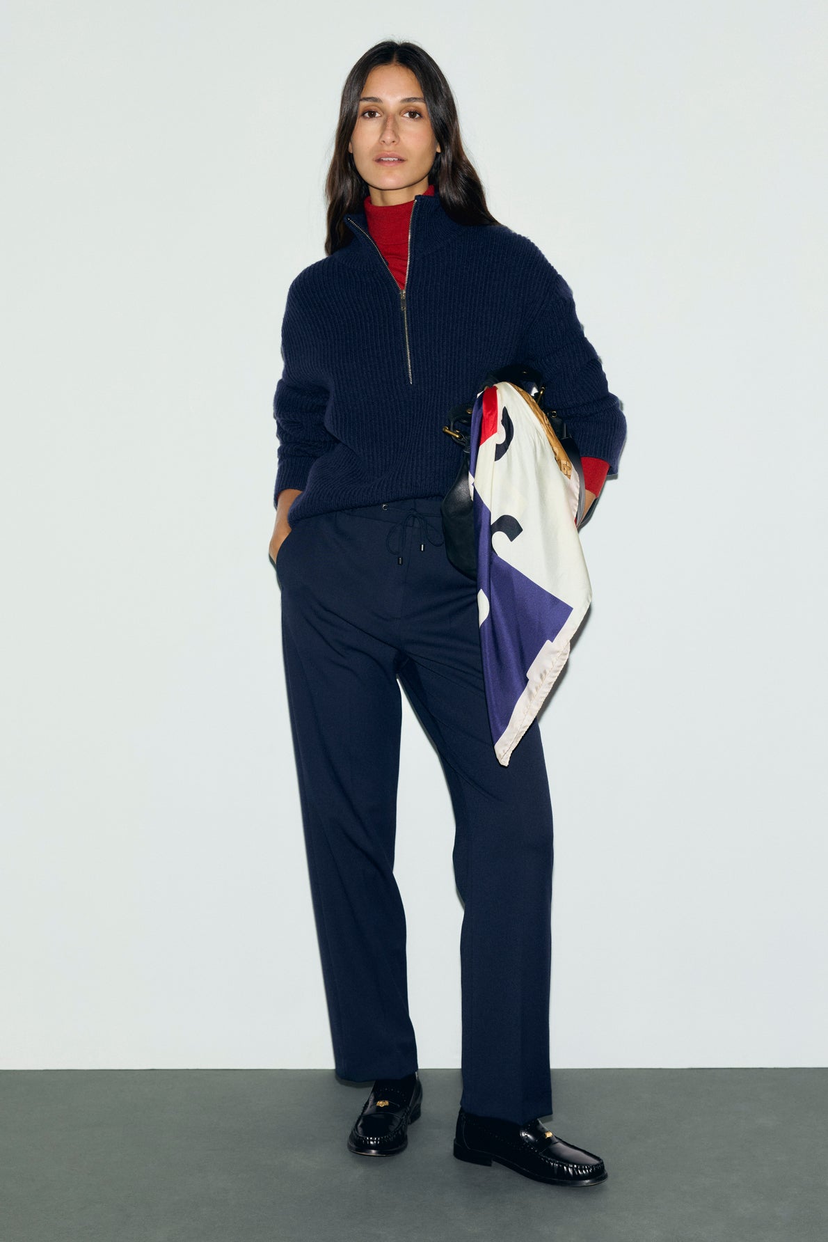 A woman stands against a plain background wearing a navy sweater, red turtleneck, pantalon bleu marine droit paired with black loafers, holding a colorful scarf draped over her arm for a casual chic look.