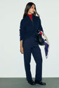 A woman stands against a light wall wearing a navy sweater, a pantalon droit bleu marine, a red turtleneck, and black loafers. She holds a black bag with a colorful scarf draped over it, showcasing an effortless pantalon casual chic style.