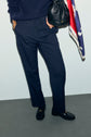 A person wearing a pantalon bleu marine, a navy sweater, and black loafers stands against a light wall, holding a black purse and a red, white, and blue scarf.