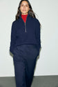 Woman standing against a plain light background, wearing a navy half-zip sweater over a red shirt and a pantalon bleu marine casual chic, with hands in her pockets and long dark hair down.