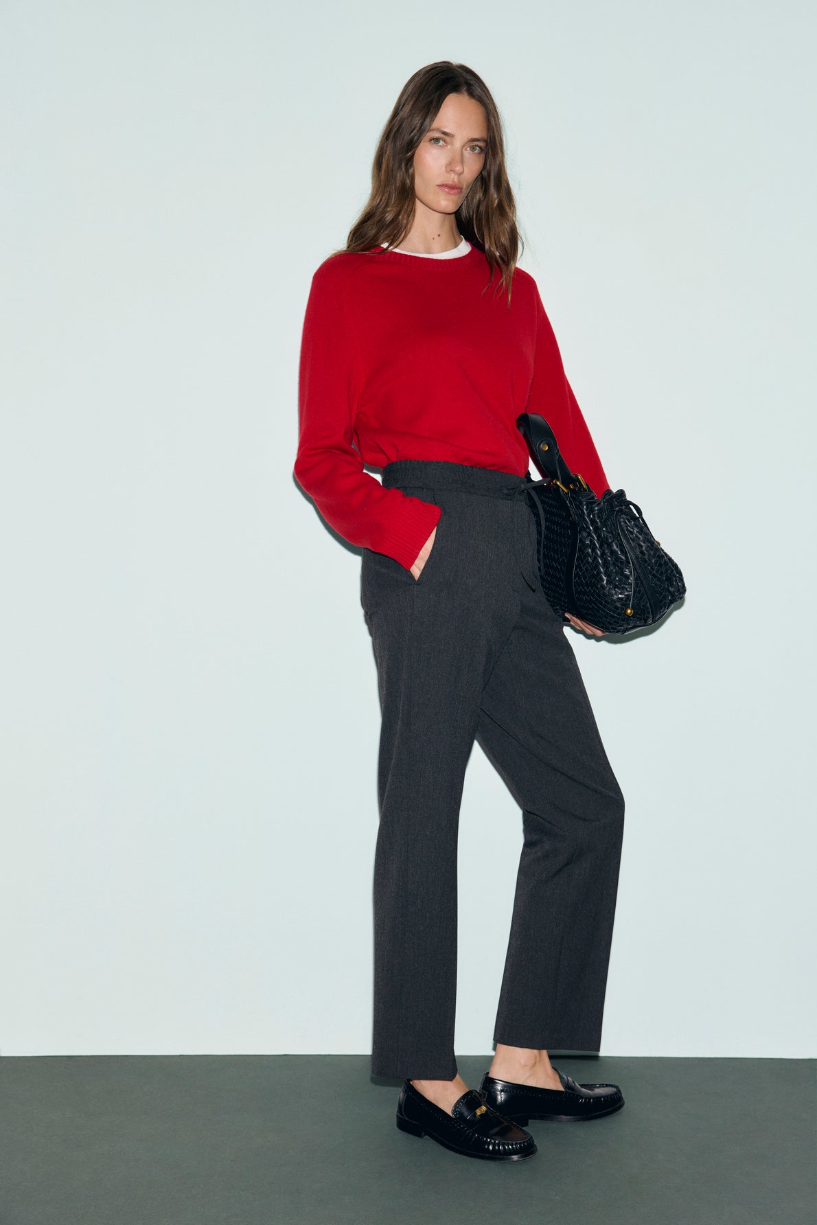 A woman stands against a plain background, wearing a red sweater and a pantalon droit gris à coupe droite. She pairs the look with black loafers and holds a large black textured handbag. Her long, wavy brown hair is styled effortlessly, completing this chic ensemble with subtle elegance.