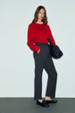 A woman stands against a plain background, wearing a red sweater and a pantalon droit gris à coupe droite. She pairs the look with black loafers and holds a large black textured handbag. Her long, wavy brown hair is styled effortlessly, completing this chic ensemble with subtle elegance.