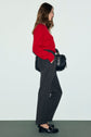 Woman in a red sweater, gray pantalon droit with dos élastiqué, and black loafers stands in profile, holding a black woven handbag against a plain light background.