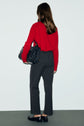 A person with long brown hair, wearing a red sweater, a pantalon droit in gray with a coupe droite fit, and black shoes, stands facing away while holding a large black handbag.