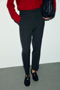 A person wearing a red sweater, dark gray pantalon droit à coupe droite with a dos élastiqué, and black loafers stands against a light wall, holding a textured black bag. Their face is not visible.